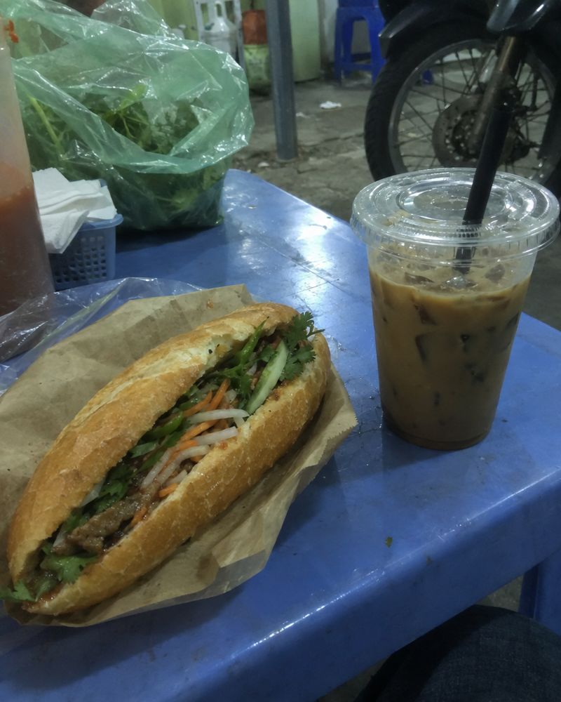 Ảnh gốc chụp vội bằng điện thoại: bánh mì và cà phê trên bàn nhựa xanh, đèn huỳnh quang, nền bừa bộn
